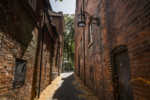 View of a narrow alleyway lined with weathered red brick walls featuring an old fashioned street lamp and a cobblestone path covered in fallen leaves Coventry England United Kingdom.