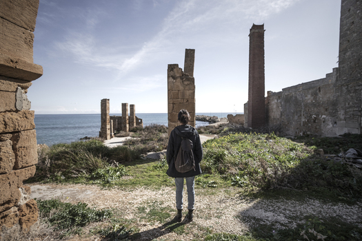 View of a woman standing among the historic stone ruins and tall chimney of the Tonnara di Avola overlooking the Mediterranean Sea in Avola Sicily Italy. View of a woman standing among the historic stone ruins and tall chimney of the Tonnara di Avola overlooking the Mediterranean Sea in Avola Sicily Italy.