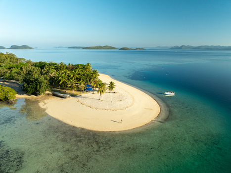 Aerial view of Pass Island featuring a white sand beach lush palm trees and clear turquoise waters with a small boat anchored offshore in Coron Philippines. Aerial view of Pass Island featuring a white sand beach lush palm trees and clear turquoise waters with a small boat anchored offshore in Coron Philippines.