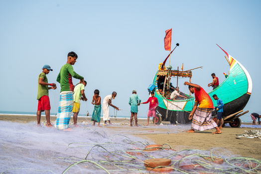 Teknaf Bangladesh 10 March 2026 View of a vibrant beach scene where fishermen mend nets under a clear sky their colorful clothing contrasting with the sandy shore.