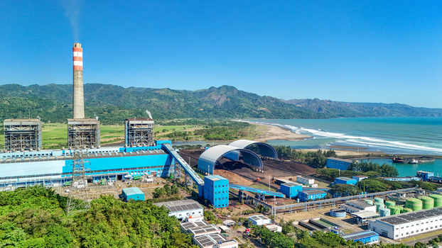 Aerial view of a power plant with large coal storage domes industrial buildings and a tall chimney situated along the coastline Indonesia.