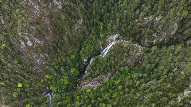 Aerial view of Salto de Aguas Blancas waterfall cascading through a lush pine forest with the winding Carretera al Salto de Aguas Blancas visible below in Constanza La Vega Dominican Republic.