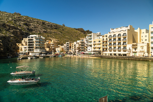 View of Xlendi Bay with turquoise water small boats moored near the shore and waterfront buildings under a clear blue sky in Zebbug Gozo Region Malta.