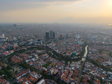 Surabaya Indonesia 28 August 2026 Aerial view of the sprawling cityscape featuring dense residential rooftops modern high rise buildings and a winding river under a soft sunset sky.