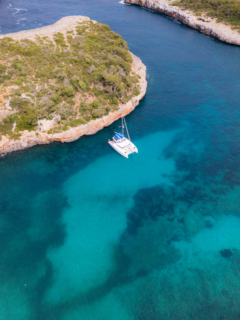 Aerial view of a white catamaran anchored in the turquoise waters of Cala Sa Nau surrounded by rocky cliffs and lush greenery Felanitx Balearic Islands Spain. Aerial view of a white catamaran anchored in the turquoise waters of Cala Sa Nau surrounded by rocky cliffs and lush greenery Felanitx Balearic Islands Spain.