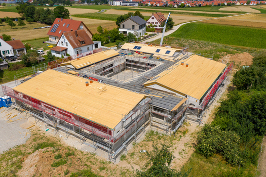 Igensdorf Germany 11 May 2024 Aerial view of a large building under construction with wooden roof panels and scaffolding surrounded by residential houses and green fields. Igensdorf Germany 11 May 2024 Aerial view of a large building under construction with wooden roof panels and scaffolding surrounded by residential houses and green fields.