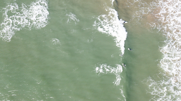 Aerial view of surfers waiting for waves in the turquoise ocean with white foam from breaking waves at Bantham Beach Kingsbridge Devon United Kingdom. Aerial view of surfers waiting for waves in the turquoise ocean with white foam from breaking waves at Bantham Beach Kingsbridge Devon United Kingdom.