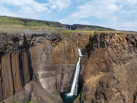 Aerial view of Litlanesfoss waterfall cascading through a dramatic canyon of basalt columns with lush green slopes under a bright blue sky in Iceland. Aerial view of Litlanesfoss waterfall cascading through a dramatic canyon of basalt columns with lush green slopes under a bright blue sky in Iceland.