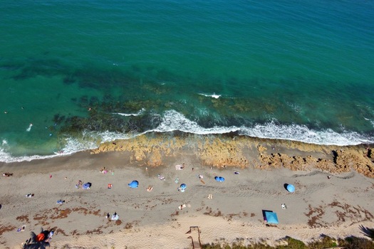 Aerial view of turquoise ocean waves crashing against a rocky shoreline with people relaxing on the sandy beach under colorful umbrellas in Jupiter Florida United States. Aerial view of turquoise ocean waves crashing against a rocky shoreline with people relaxing on the sandy beach under colorful umbrellas in Jupiter Florida United States.