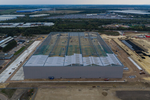 Aerial view of a large industrial warehouse under construction with a steel frame structure and metal roofing panels situated in a vast development site in the United Kingdom. Aerial view of a large industrial warehouse under construction with a steel frame structure and metal roofing panels situated in a vast development site in the United Kingdom.