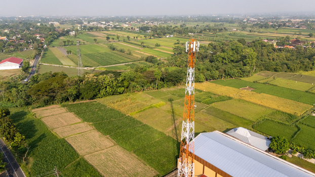 Aerial view of a telecommunications tower standing amidst lush green agricultural fields and rice paddies in a rural landscape in Indonesia.