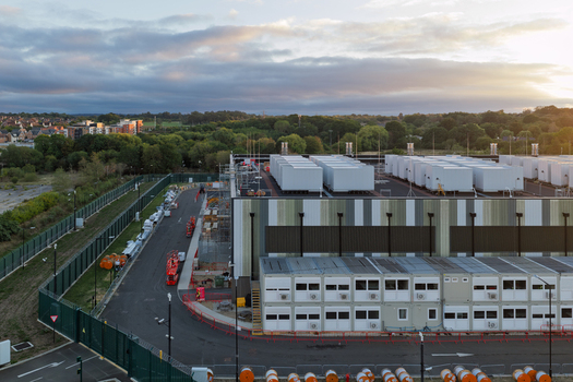 Aerial view of a large data center facility with modular office units and construction equipment surrounded by a security fence and lush green trees in the United Kingdom. Aerial view of a large data center facility with modular office units and construction equipment surrounded by a security fence and lush green trees in the United Kingdom.