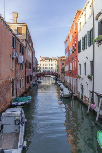 View of a narrow canal lined with historic colorful buildings a wooden footbridge and moored boats under a clear sky in Venice Veneto Italy. View of a narrow canal lined with historic colorful buildings a wooden footbridge and moored boats under a clear sky in Venice Veneto Italy.