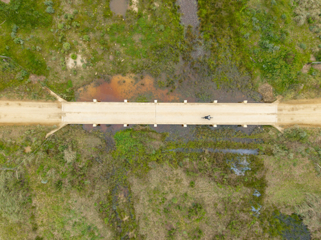 Aerial view of a cyclist riding across a narrow dirt road bridge over a marshy wetland area with lush green vegetation and small pools of water in Greyton Western Cape South Africa.