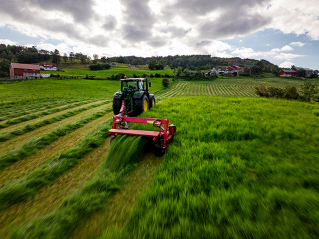Aerial view of a tractor performing silage production in a lush green field with farmhouses in the background in Rogaland Norway.