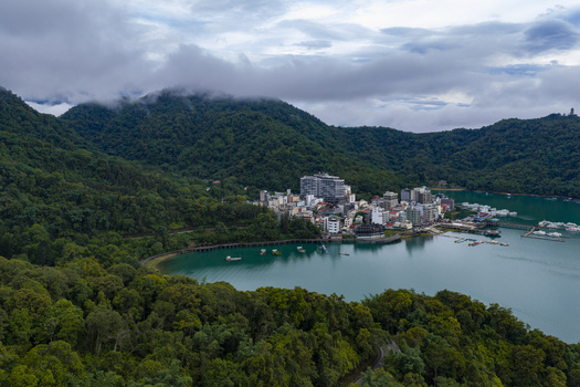 Aerial view of Sun Moon Lake surrounded by lush green mountains hotels and a pier with boats under a cloudy sky in Yuchi Township Nantou County Taiwan. Aerial view of Sun Moon Lake surrounded by lush green mountains hotels and a pier with boats under a cloudy sky in Yuchi Township Nantou County Taiwan.