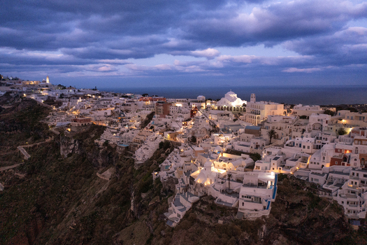 Aerial view of the iconic white washed buildings and a majestic domed church glowing softly against the dramatic dark cliffs and twilight sky Fira Thira Greece. Aerial view of the iconic white washed buildings and a majestic domed church glowing softly against the dramatic dark cliffs and twilight sky Fira Thira Greece.