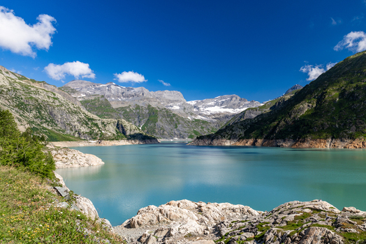 View of the turquoise waters of Emosson Lake at VerticAlp Emosson surrounded by rocky mountain slopes and snow capped peaks under a clear blue sky in Le Chatelard Fribourg Switzerland. View of the turquoise waters of Emosson Lake at VerticAlp Emosson surrounded by rocky mountain slopes and snow capped peaks under a clear blue sky in Le Chatelard Fribourg Switzerland.