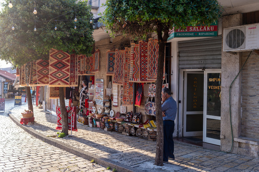 Kruje Albania 12 June 2026 View of a traditional bazaar shop displaying colorful woven rugs and souvenirs along a cobblestone street with a man standing near a tree.