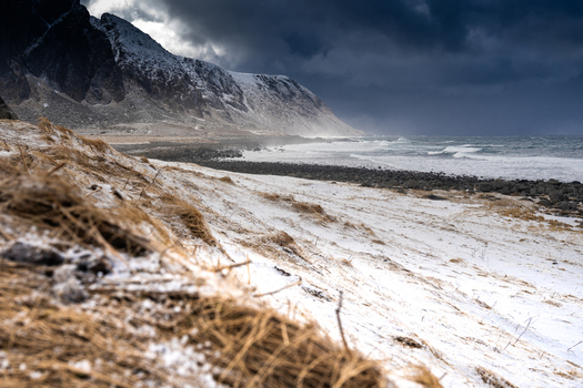 View of a snow covered beach with dry grass in the foreground dramatic dark storm clouds over the mountains and waves crashing on the rocky shore in Leknes Lofoten Norway.