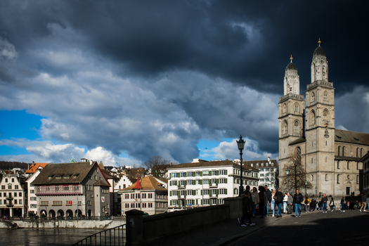 Zurich Switzerland 02 April 2023 View of the Grossmünster church standing tall against a dramatic sky as people stroll along the Limmat river its waters reflecting the city s architectural charm. Zurich Switzerland 02 April 2023 View of the Grossmünster church standing tall against a dramatic sky as people stroll along the Limmat river its waters reflecting the city s architectural charm.