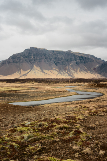 View of a winding river flowing through a mossy landscape towards a large mountain range under a cloudy sky in Borgarbyggd Iceland. View of a winding river flowing through a mossy landscape towards a large mountain range under a cloudy sky in Borgarbyggd Iceland.