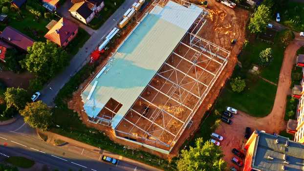 Aerial view of a construction site featuring a steel frame structure and partially installed metal roof surrounded by residential houses roads and greenery in Latvija. Aerial view of a construction site featuring a steel frame structure and partially installed metal roof surrounded by residential houses roads and greenery in Latvija.
