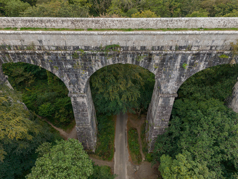 Aerial view of the historic stone Treffry Viaduct spanning a lush green valley with a narrow road passing beneath its arches in Luxulyan Cornwall United Kingdom.