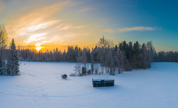 Aerial view of a barrel sauna and a small cabin in a snow covered field surrounded by a forest at sunset in Latvia. Aerial view of a barrel sauna and a small cabin in a snow covered field surrounded by a forest at sunset in Latvia.