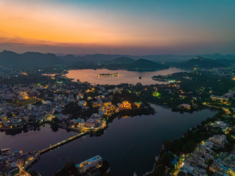 Aerial view of Lake Pichola and the illuminated City Palace at dusk with surrounding historic architecture calm waters and distant Aravalli hills in Udaipur Rajasthan India.