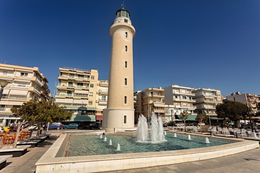 View of the historic Alexandroupoli Lighthouse standing tall next to a fountain in a city square with surrounding buildings under a clear blue sky in Alexandroupoli Greece.