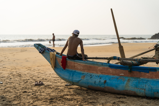 Sri Lanka 16 January 2026 View of a young man sitting on a traditional blue wooden boat on a sandy beach with ocean waves in the background.