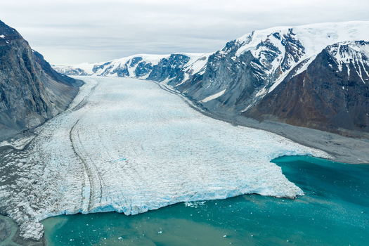 Aerial view of a massive glacier flowing between rugged snow capped mountains into a turquoise glacial lake in Greenland.