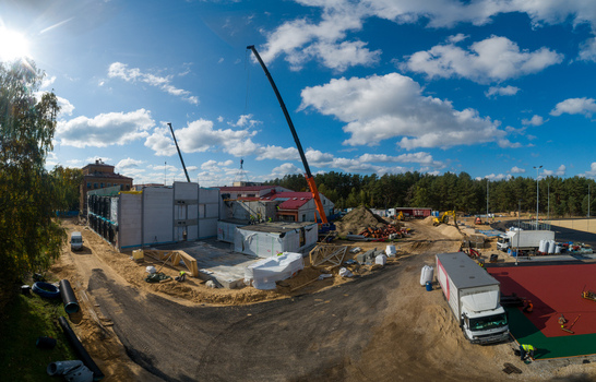 Aerial view of a construction site featuring cranes building structures a red sports surface and trucks under a blue sky in Latvia. Aerial view of a construction site featuring cranes building structures a red sports surface and trucks under a blue sky in Latvia.
