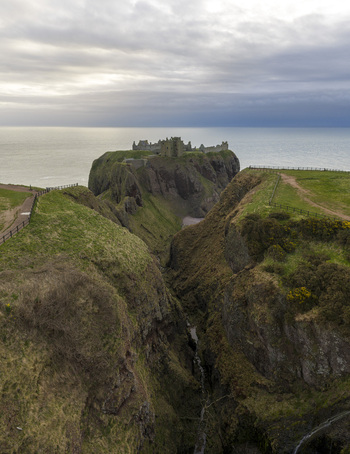 Aerial view of Dunnottar Castle perched on a rocky headland overlooking the North Sea with steep cliffs and green hills under an overcast sky Stonehaven Scotland United Kingdom. Aerial view of Dunnottar Castle perched on a rocky headland overlooking the North Sea with steep cliffs and green hills under an overcast sky Stonehaven Scotland United Kingdom.