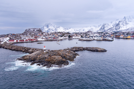 Aerial view of the Lofoten islands featuring a lighthouse on a rocky outcrop traditional red houses and snow covered mountains in the background in Nordland Norway.