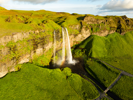 Aerial view of Seljalandsfoss waterfall cascading over rocky cliffs into a lush green landscape with a visible rainbow at the base and walking paths below in Rangarthing eystra Iceland. Aerial view of Seljalandsfoss waterfall cascading over rocky cliffs into a lush green landscape with a visible rainbow at the base and walking paths below in Rangarthing eystra Iceland.