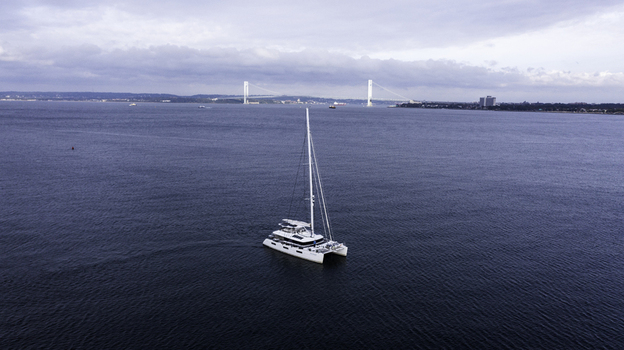 Aerial view of a white catamaran sailing on the calm blue waters of the bay with the Verrazzano Narrows Bridge in the background in Kings County New York United States. Aerial view of a white catamaran sailing on the calm blue waters of the bay with the Verrazzano Narrows Bridge in the background in Kings County New York United States.