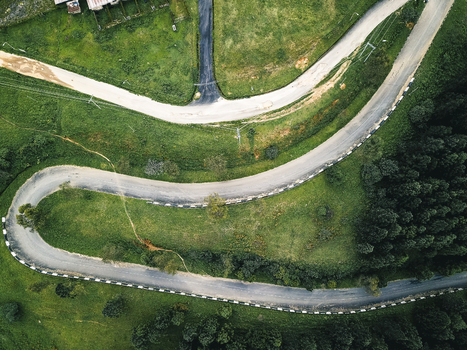 Aerial view of a winding asphalt road cutting through vibrant green hills and dark forest with a lighter dirt path branching off Obudu Cross River Nigeria.