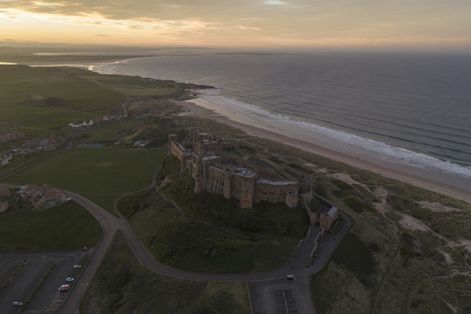 Aerial view of Bamburgh Castle overlooking the sandy beach and North Sea under a golden sunset sky in Bamburgh England United Kingdom. Aerial view of Bamburgh Castle overlooking the sandy beach and North Sea under a golden sunset sky in Bamburgh England United Kingdom.