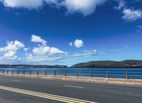 View of the coastal road and wooden fence overlooking the blue waters and green hills of Plettenberg Bay Western Cape South Africa. View of the coastal road and wooden fence overlooking the blue waters and green hills of Plettenberg Bay Western Cape South Africa.