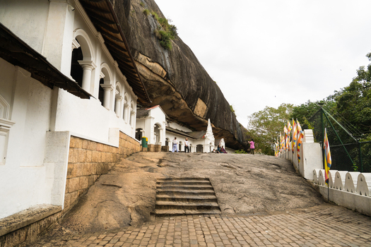 View of the historic Dambulla Cave Temple complex with white arched buildings nestled under a massive rock overhang and stone steps Dambulla Central Province Sri Lanka.