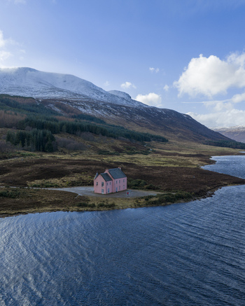 Aerial view of The Pink House on the shore of Loch Glass with snow capped mountains and a pine forest under a blue sky in Dingwall Scotland United Kingdom. Aerial view of The Pink House on the shore of Loch Glass with snow capped mountains and a pine forest under a blue sky in Dingwall Scotland United Kingdom.