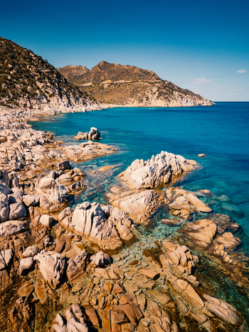 Aerial view of Punta Molentis featuring granite rock formations along the turquoise coastline and rugged hills under a clear blue sky in Villasimius Sardegna Italy.