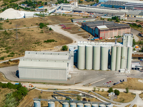 Aerial view of an industrial facility featuring large cylindrical grain silos and expansive warehouse buildings surrounded by a dry landscape in Hombourg Grand Est France.