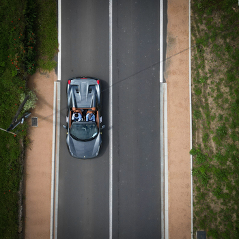 Aerial view of a grey convertible sports car with two people driving along a paved road surrounded by green grass in La Chapelle de Guinchay Bourgogne Franche Comte France.