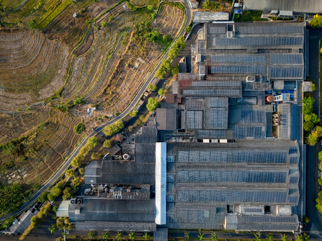 Aerial view of a large industrial factory complex with extensive solar panel arrays on the roof situated next to terraced rice fields and a road in Semarang Regency Central Java Indonesia. Aerial view of a large industrial factory complex with extensive solar panel arrays on the roof situated next to terraced rice fields and a road in Semarang Regency Central Java Indonesia.