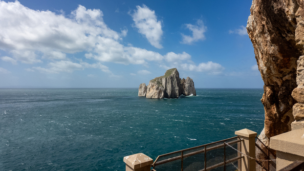 View of Scoglio Pan di zucchero sea stack rising from the Mediterranean sea under a blue sky with white clouds in Masua Sardegna Italy. View of Scoglio Pan di zucchero sea stack rising from the Mediterranean sea under a blue sky with white clouds in Masua Sardegna Italy.