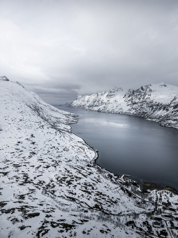 Aerial view of the deep blue waters of Ersfjorden fjord surrounded by steep snow covered mountains under a moody overcast sky in Ersfjord Troms Norway. Aerial view of the deep blue waters of Ersfjorden fjord surrounded by steep snow covered mountains under a moody overcast sky in Ersfjord Troms Norway.