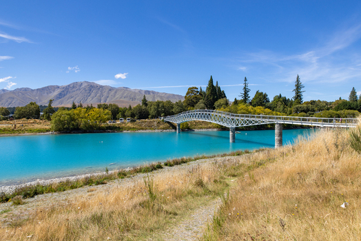 View of vibrant turquoise waters flow beneath a white bridge under a clear blue sky framed by golden grasses and distant mountains Lake Tekapo New Zealand. View of vibrant turquoise waters flow beneath a white bridge under a clear blue sky framed by golden grasses and distant mountains Lake Tekapo New Zealand.